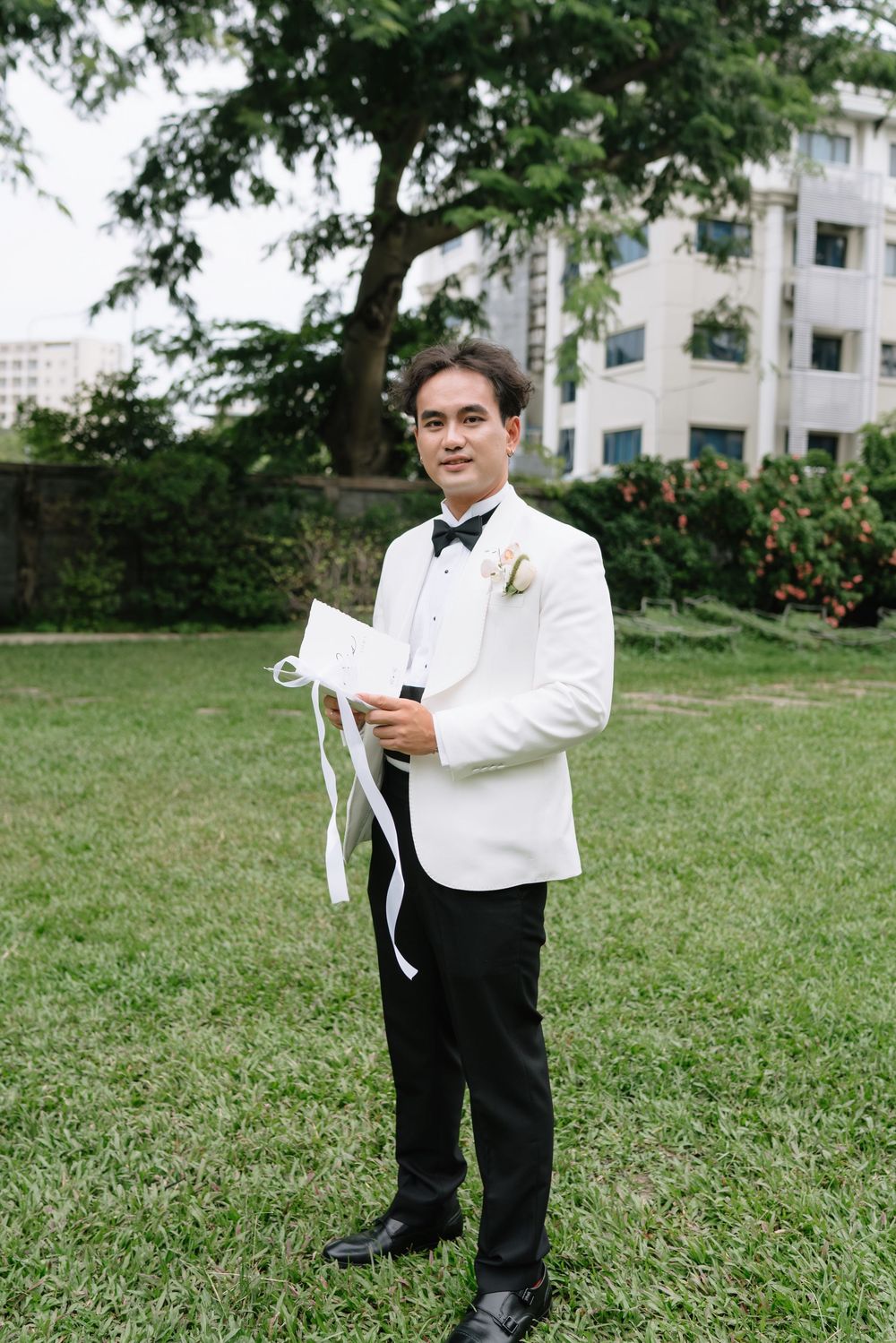 Groom in white tuxedo and black bow tie holding vow booklet while standing on green lawn.