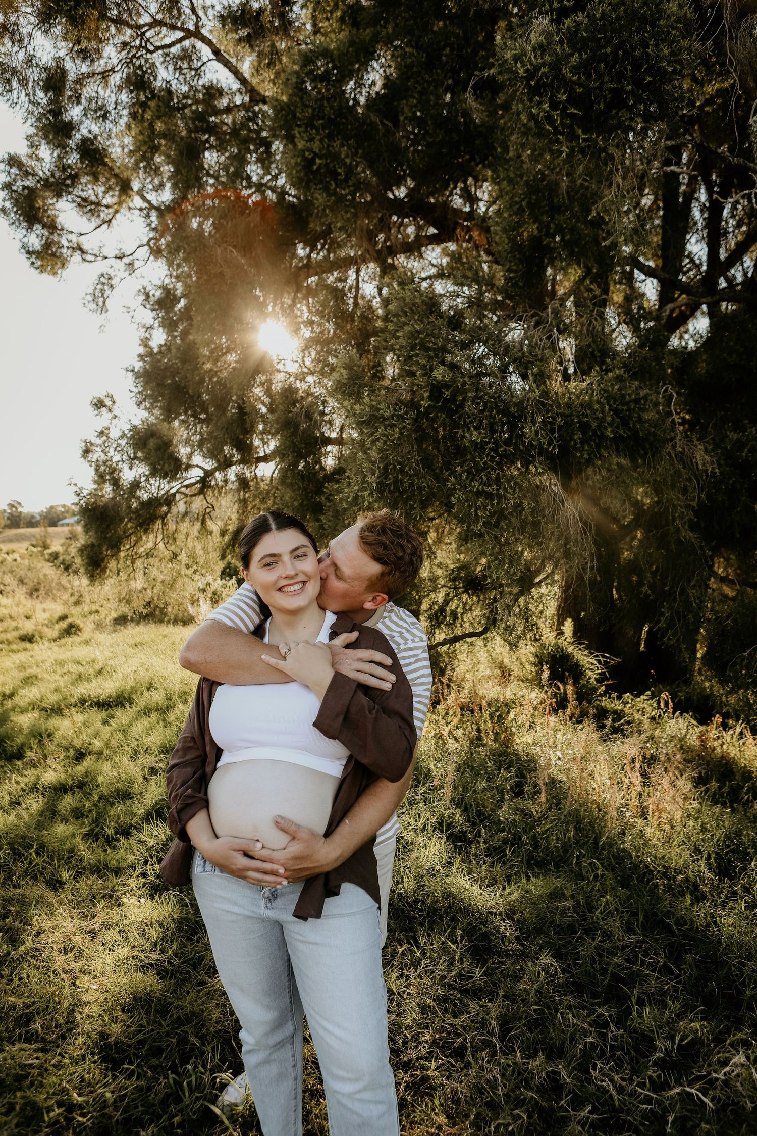 A romantic couple embraces in golden sunlight among trees during an outdoor photo session at sunset.