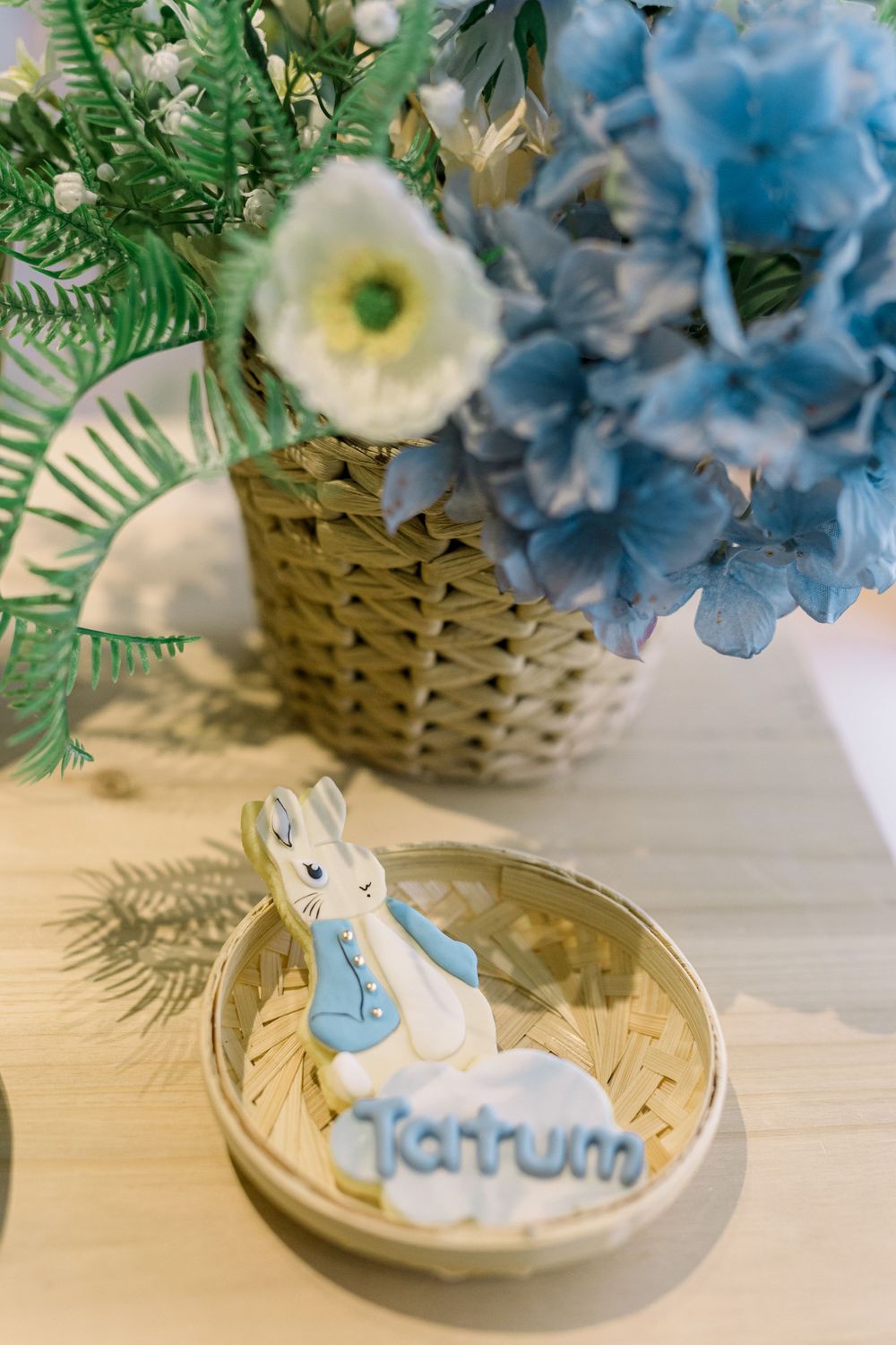 A Peter Rabbit figurine sits near a woven basket filled with blue hydrangeas and white flowers on a wooden surface.