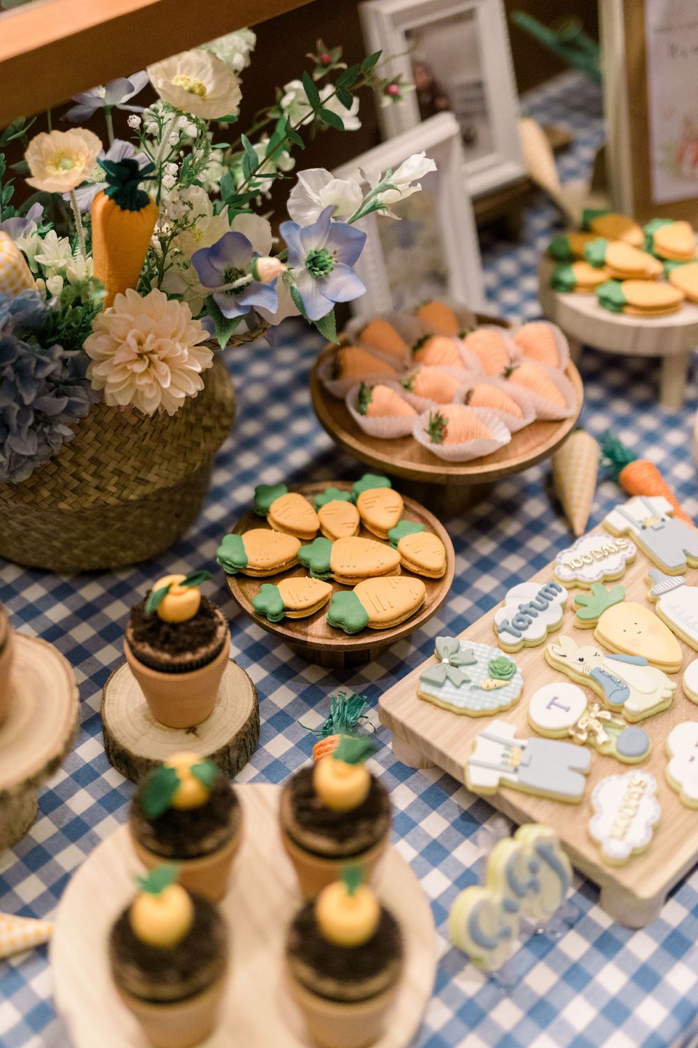 Dessert table with macarons, cookies and floral arrangements displayed on a blue checkered tablecloth.