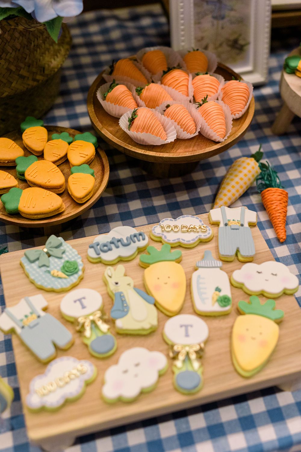 An assortment of decorated cookies featuring carrots, clouds, and baby-themed designs arranged on a blue checkered tablecloth.