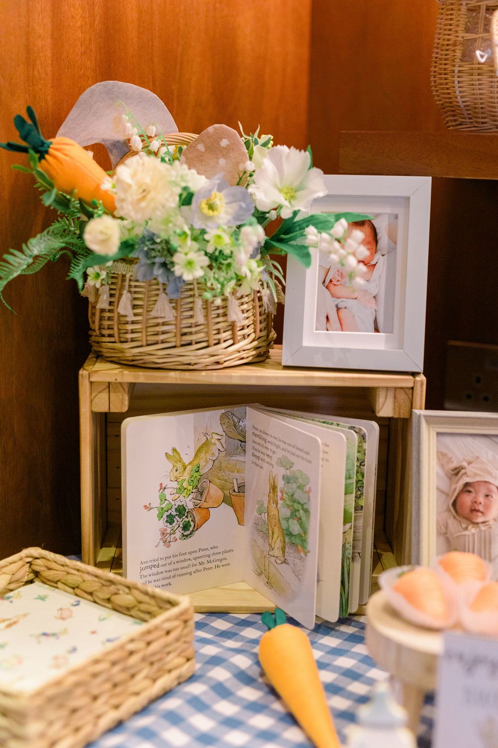 Whimsical Easter decor display with woven basket of flowers, children's book, and decorative carrots on blue gingham.
