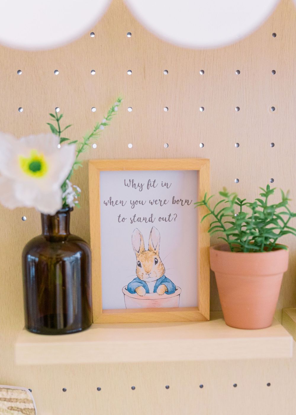 A Peter Rabbit themed artwork in wooden frame sits on a shelf with a dark bottle vase and small potted plant.