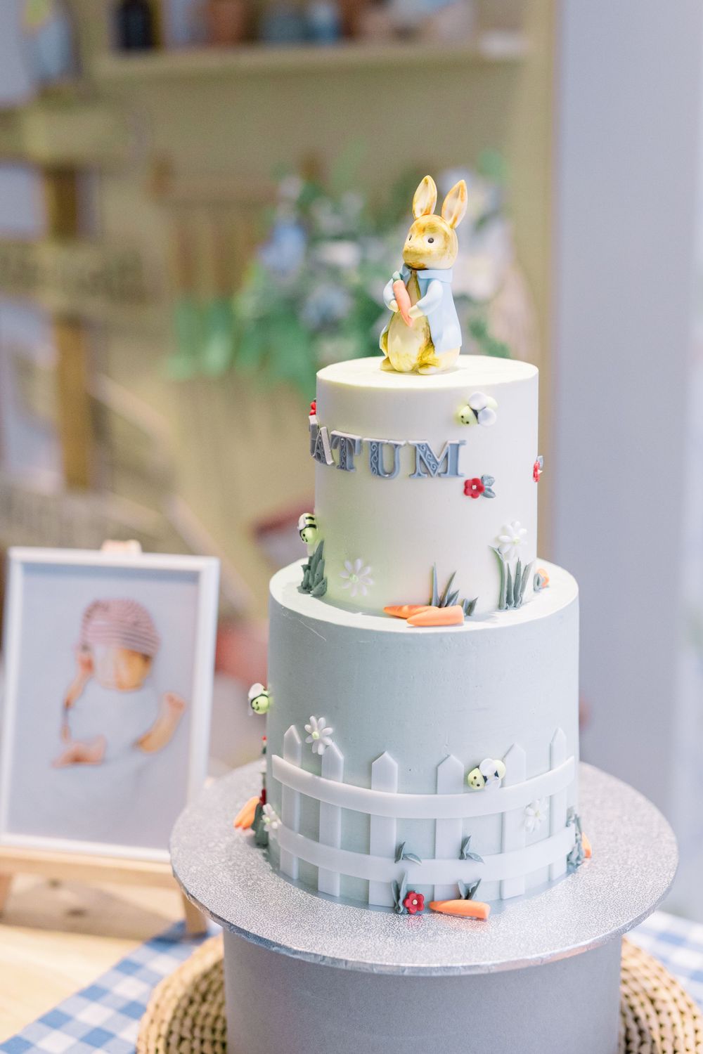 A three-tiered white and blue Peter Rabbit themed birthday cake with bunny topper and garden fence decorations.