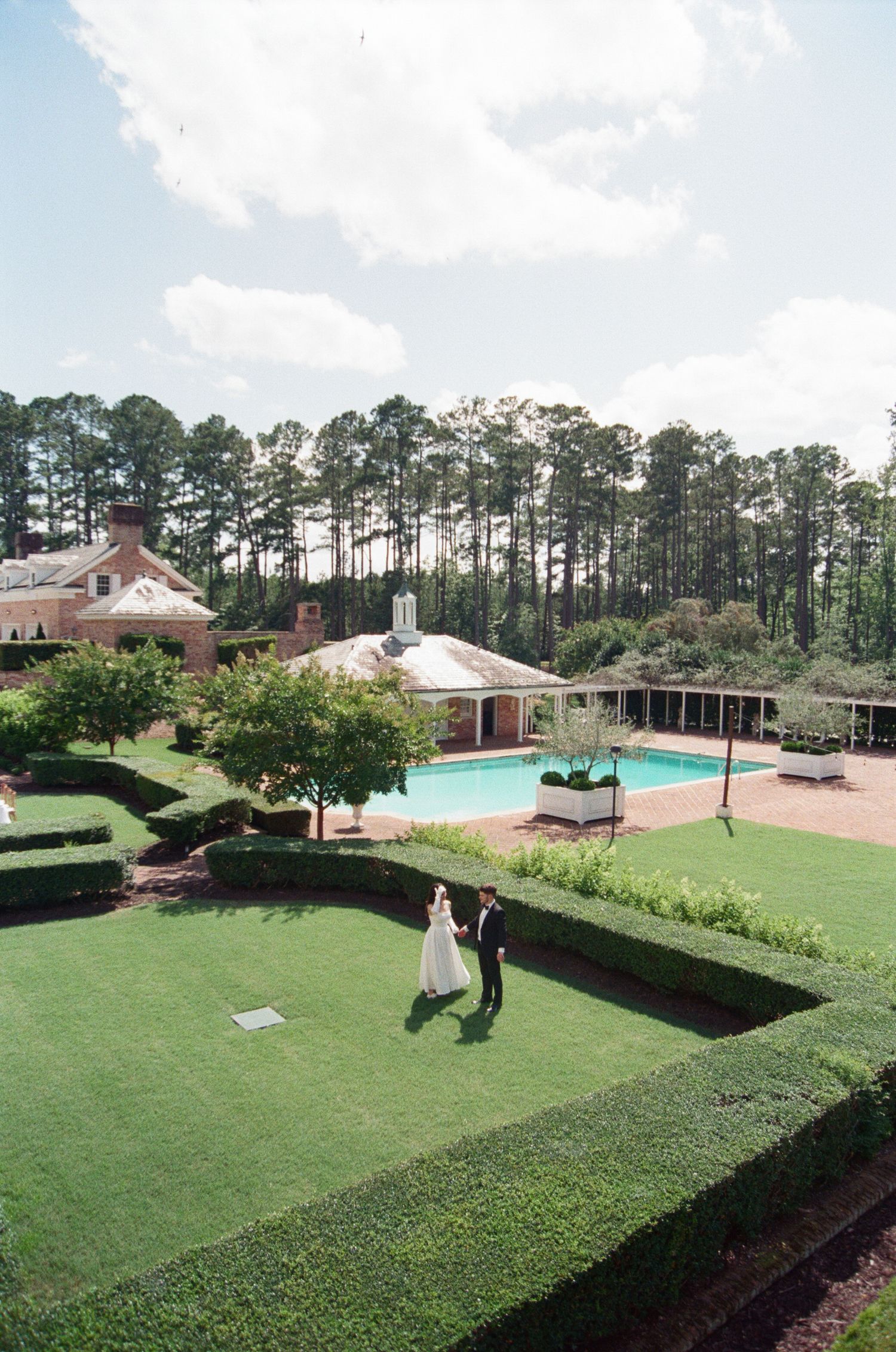 Panoramic view of an elegant outdoor venue with manicured lawns, swimming pool, and tall pine trees in the background.