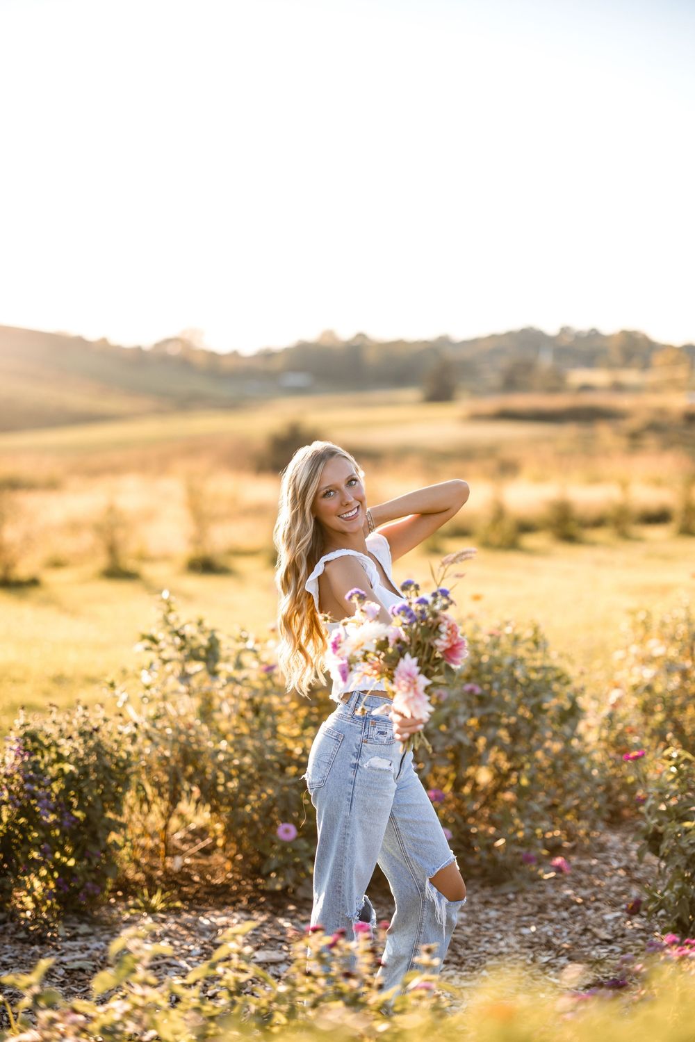 A person in denim overalls poses playfully in a golden field at sunset among wildflowers and shrubs.