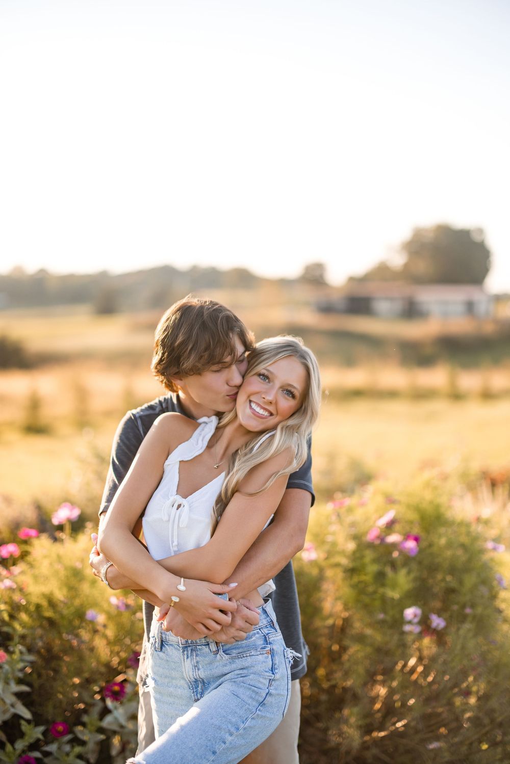 A romantic couple embraces in a golden field at sunset with pink wildflowers in the background.