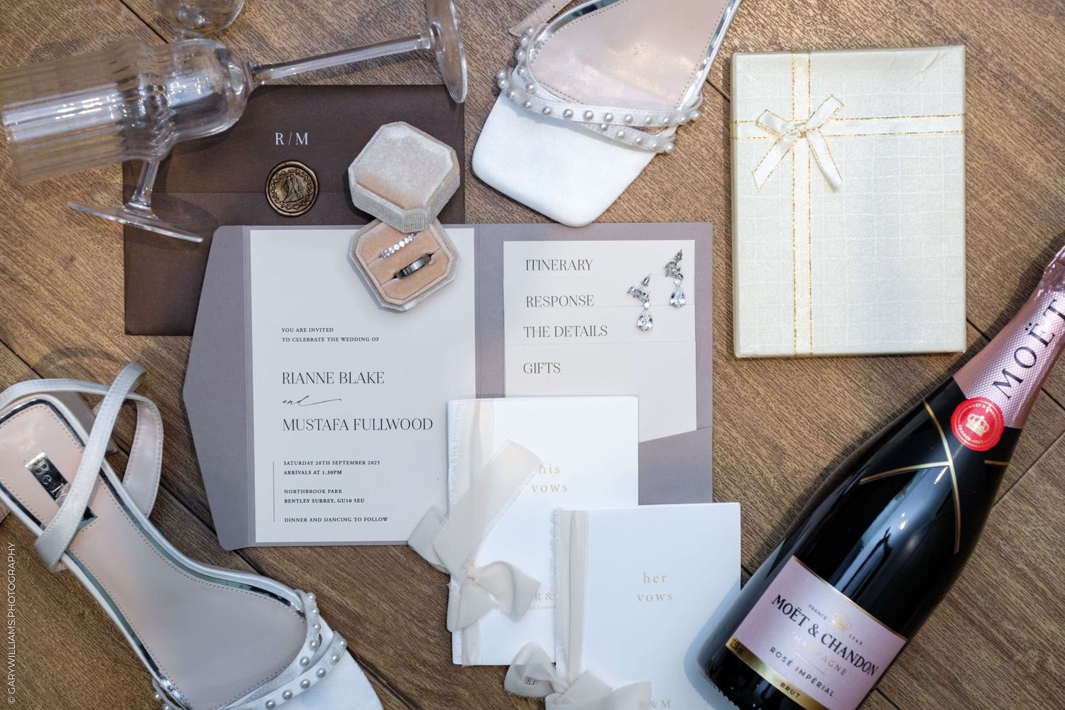 Wedding invitation suite with champagne bottle, high heel shoes and accessories arranged on herringbone wood flooring.