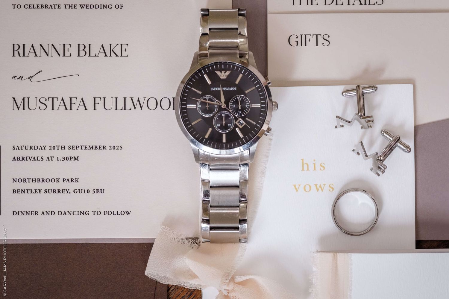 Wedding invitation details with a silver watch, cufflinks and ring arranged on white paper.
