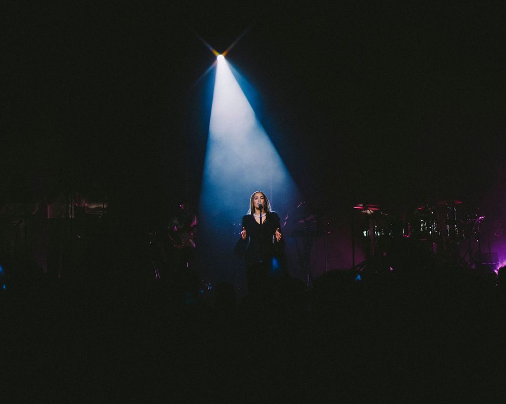 A performer stands on a dark stage illuminated by a dramatic blue spotlight during a concert performance.
