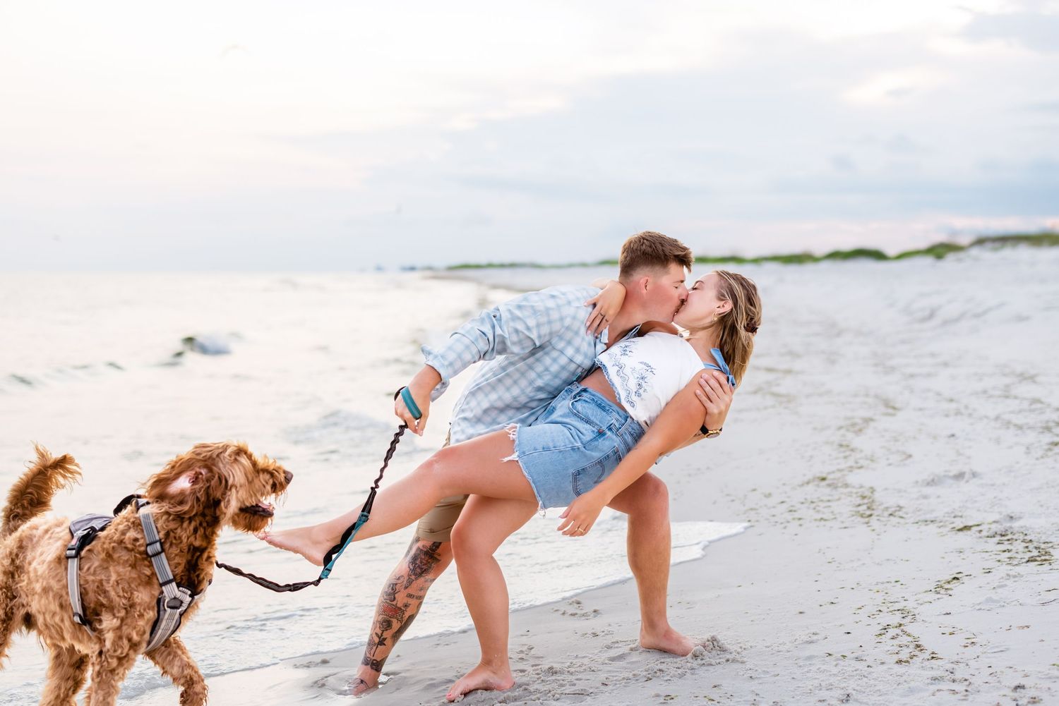 Two people and a brown dog playing and having fun together on a white sandy beach at sunset.