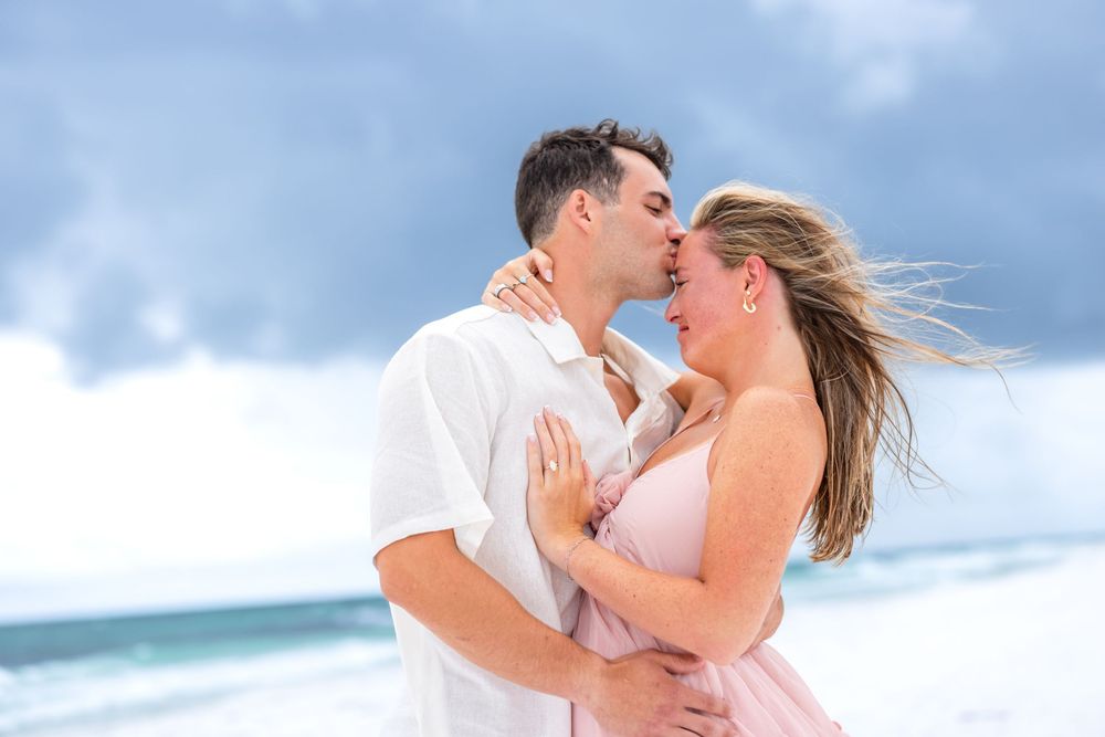 A romantic couple shares a tender moment on a breezy beach with ocean waves and clouds in the background.