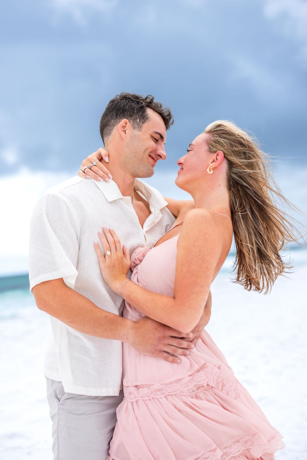 A romantic couple embraces on a breezy beach, with her pink dress flowing in the wind against a blue sky.
