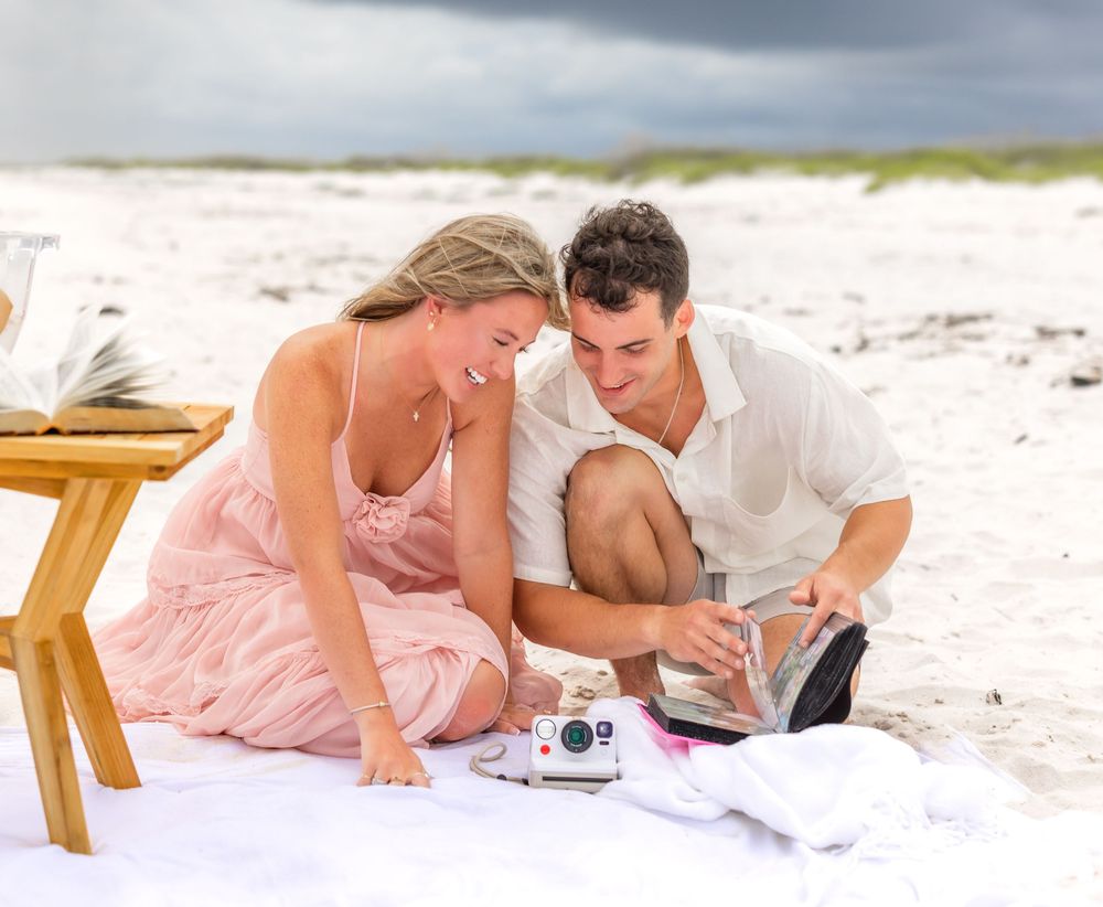 Two people relax together on a sandy white beach while looking at a digital camera.