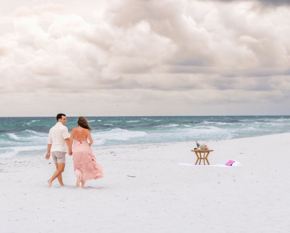 A couple in pink and white attire shares a romantic moment walking along a pristine white sandy beach by turquoise waters.