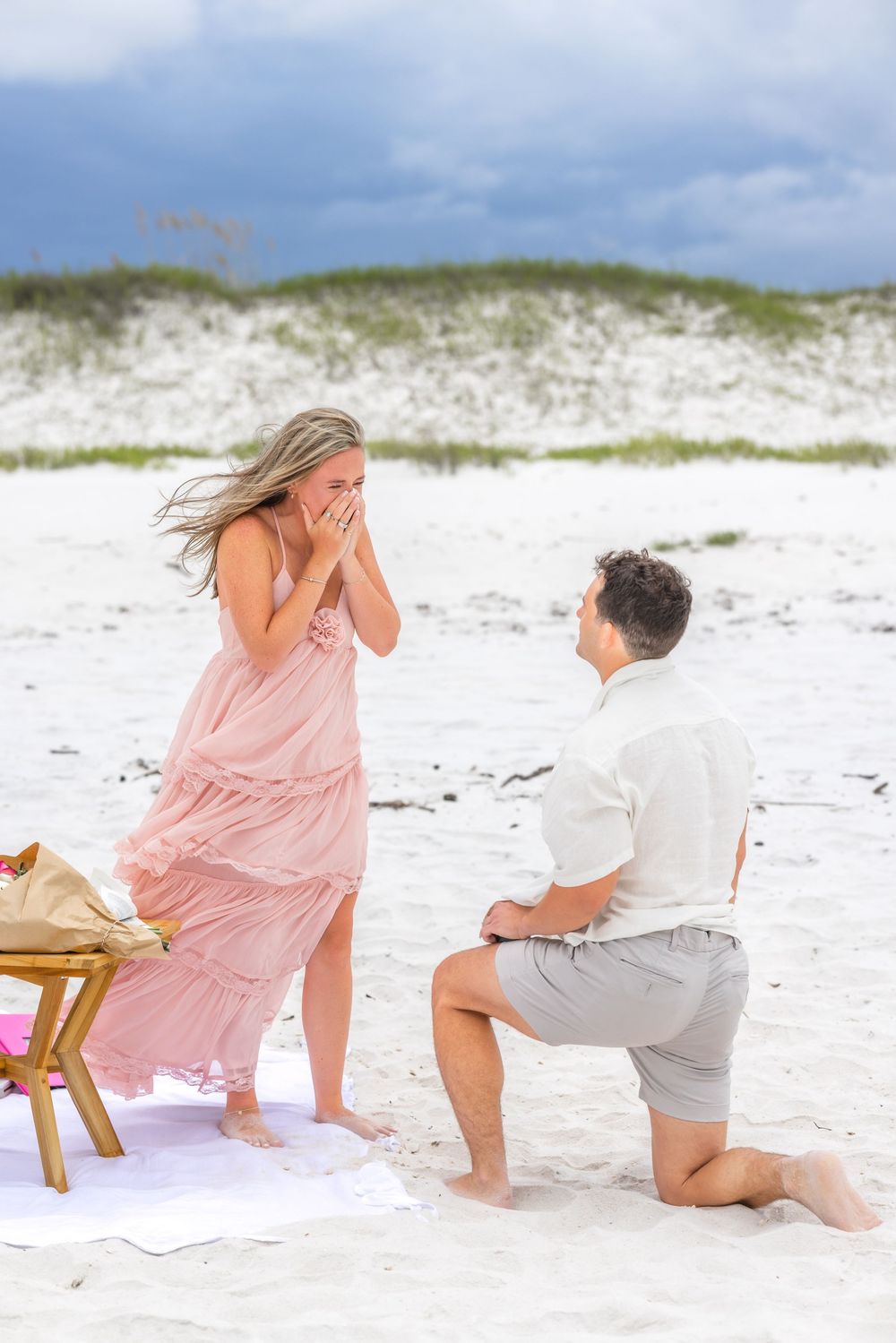 A surprise beach proposal scene unfolds with a wooden table and drinks on a white sandy beach.
