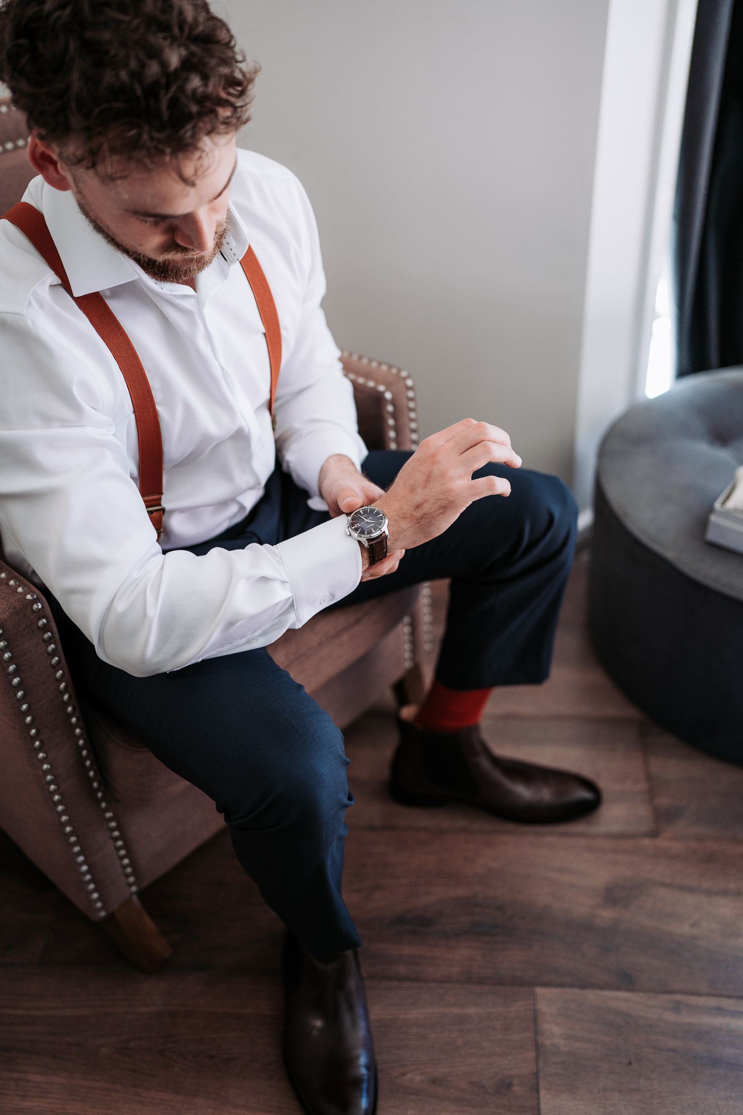 A person in formal attire with brown suspenders checks their watch while seated in a leather armchair.