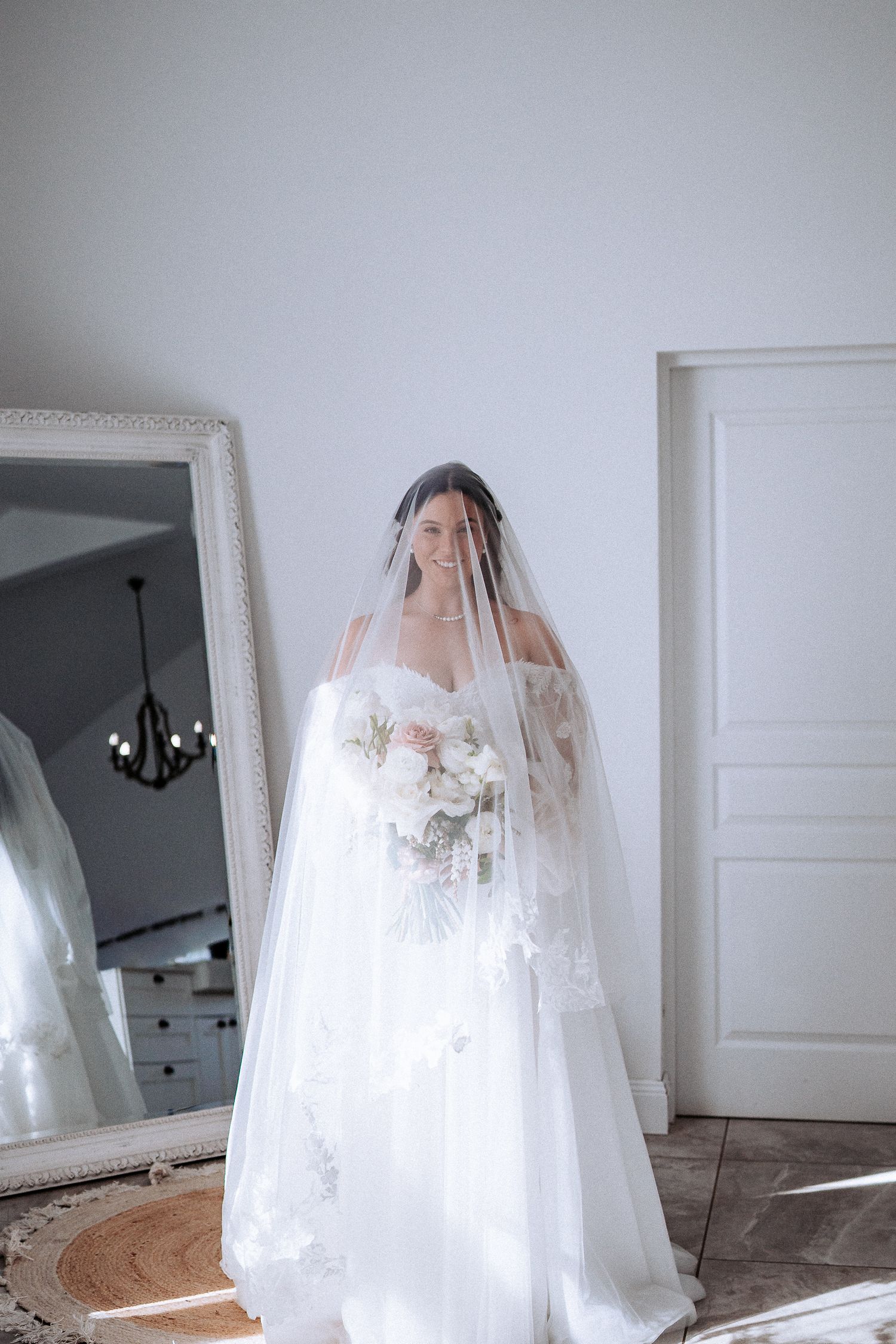 A figure in a flowing white wedding gown and veil poses in front of a mirror in a sunlit room.