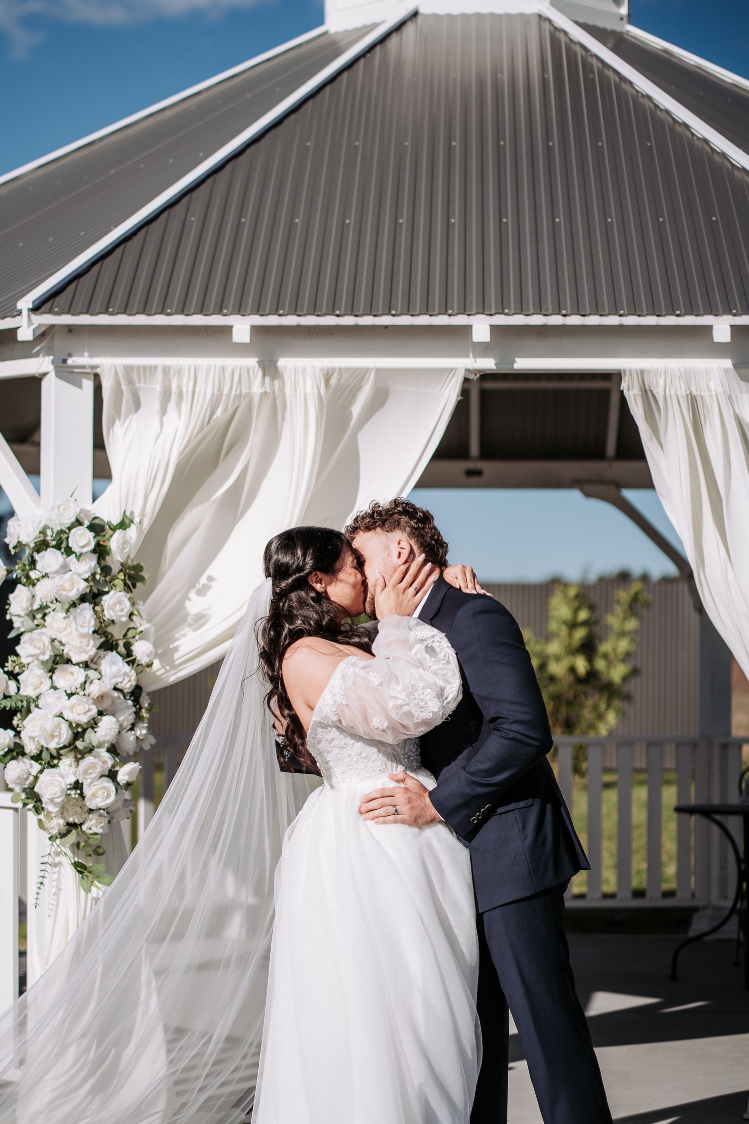 Wedding couple shares intimate kisses in a white gazebo decorated with flowing curtains and floral arrangements.