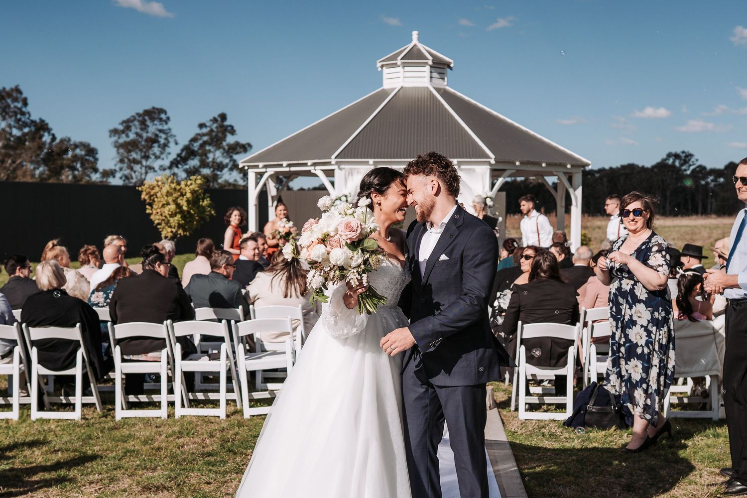 Wedding ceremony takes place in front of white gazebo at outdoor venue on sunny day.