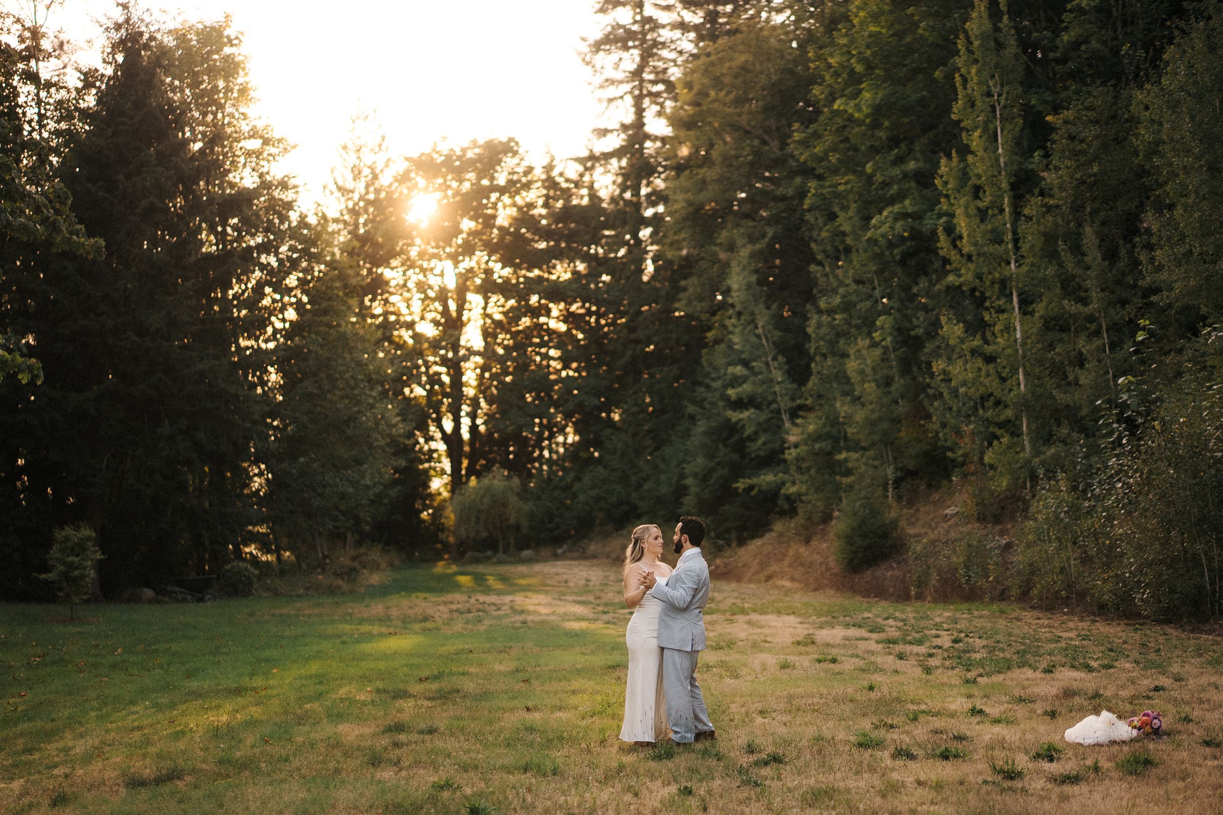 A couple dances together in a grassy meadow at sunset with tall evergreen trees in the background.