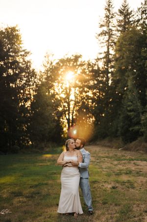 A loving couple shares an intimate moment in a sunlit meadow at golden hour with evergreen trees in the background.