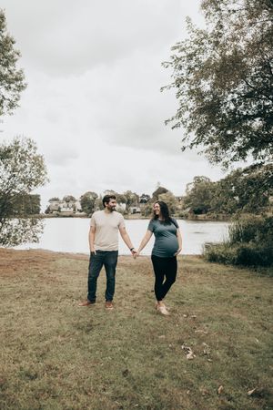 A couple walks hand in hand on a grassy hillside overlooking a serene lake on an overcast fall day.