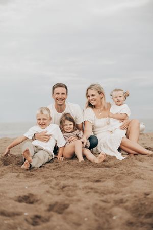 A family in white clothing sits playfully together on a sandy beach against an overcast sky.