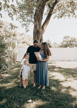 A family walks together in a sunlit garden setting, with trees providing gentle shade on a warm day.