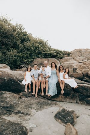 Family wearing coordinated white outfits relaxing on coastal rocks in beautiful natural lighting.