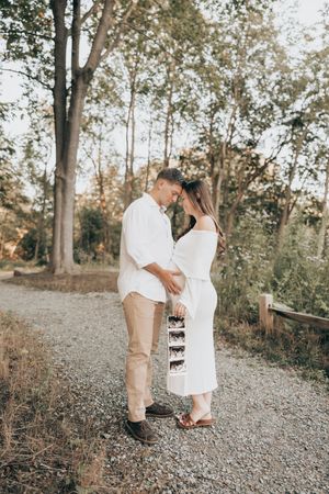 Couple in white and beige outfits standing on gravel path amid autumn trees during golden hour session.