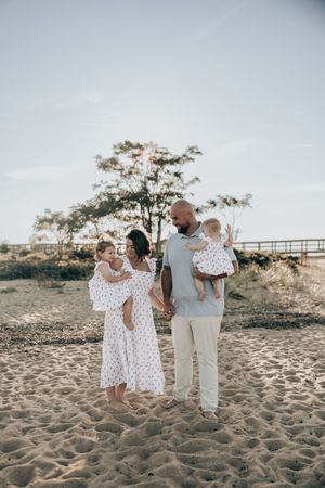 A family walks together on a sandy beach during a sunset photoshoot in casual outfits.