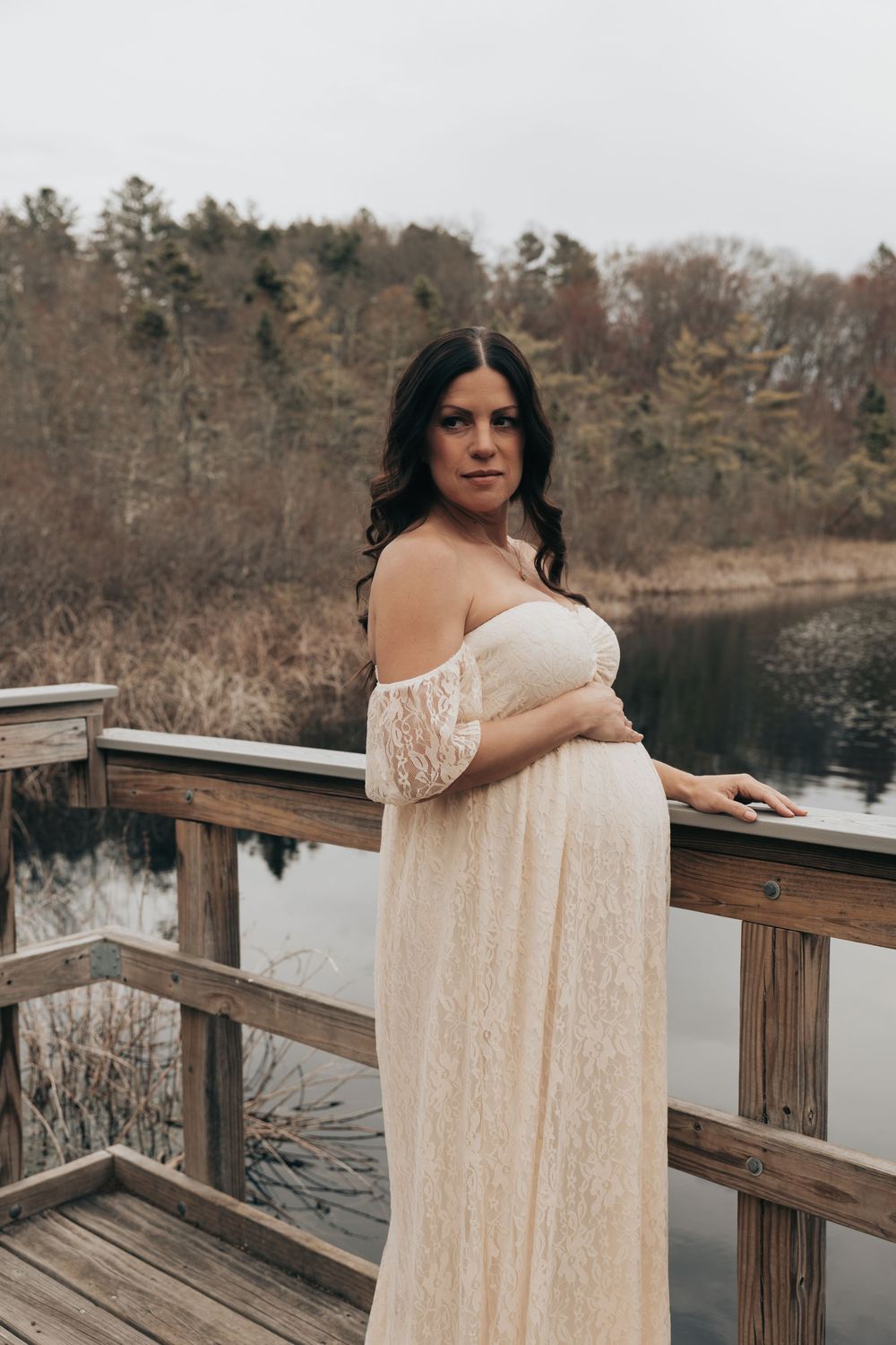 A person in a flowing cream lace dress stands contemplatively on a wooden deck overlooking a serene lake.