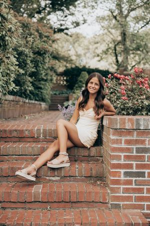 A woman in a cream-colored dress sits on brick stairs next to blooming flowers in a garden setting.