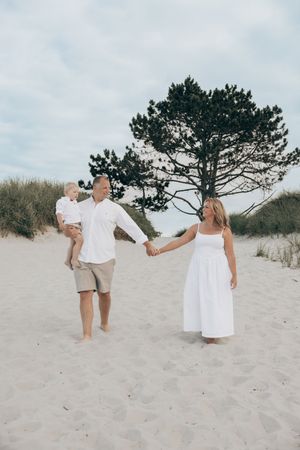 A couple in white and beige outfits walking and dancing together on a sandy beach near pine trees.