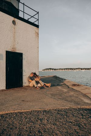 Couple resting together on a concrete pier at sunset overlooking the ocean against a white lighthouse wall.