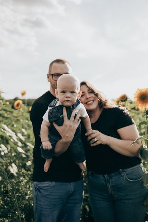 Happy couple in black shirts laughing together with their baby in a sunflower field during outdoor photo session.