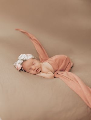 Sleeping newborn in peachy-pink wrap and white headband on neutral beige backdrop in professional studio setting.