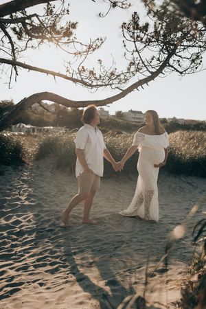 A couple walks hand in hand along a sandy beach path during golden hour with sunlight filtering through trees.