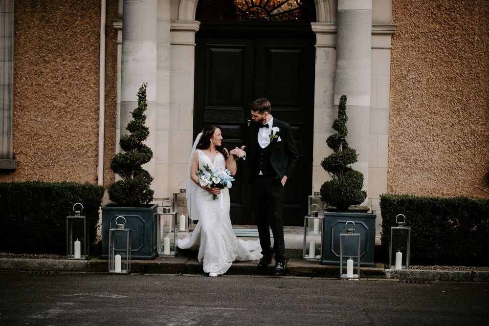A couple exits an elegant church doorway with topiary trees and lanterns framing the entrance on their wedding day.