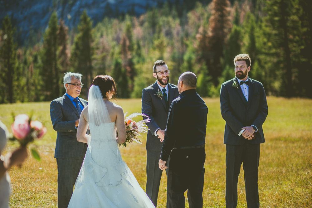 Wedding party celebrates outdoors in a sunny meadow with mountains and evergreen trees in the background. Banff Micro Wedding Photographer