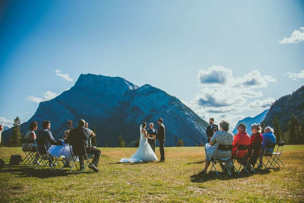 An outdoor wedding ceremony takes place in a grassy meadow with majestic mountain peaks and blue skies in Banff, Canada.