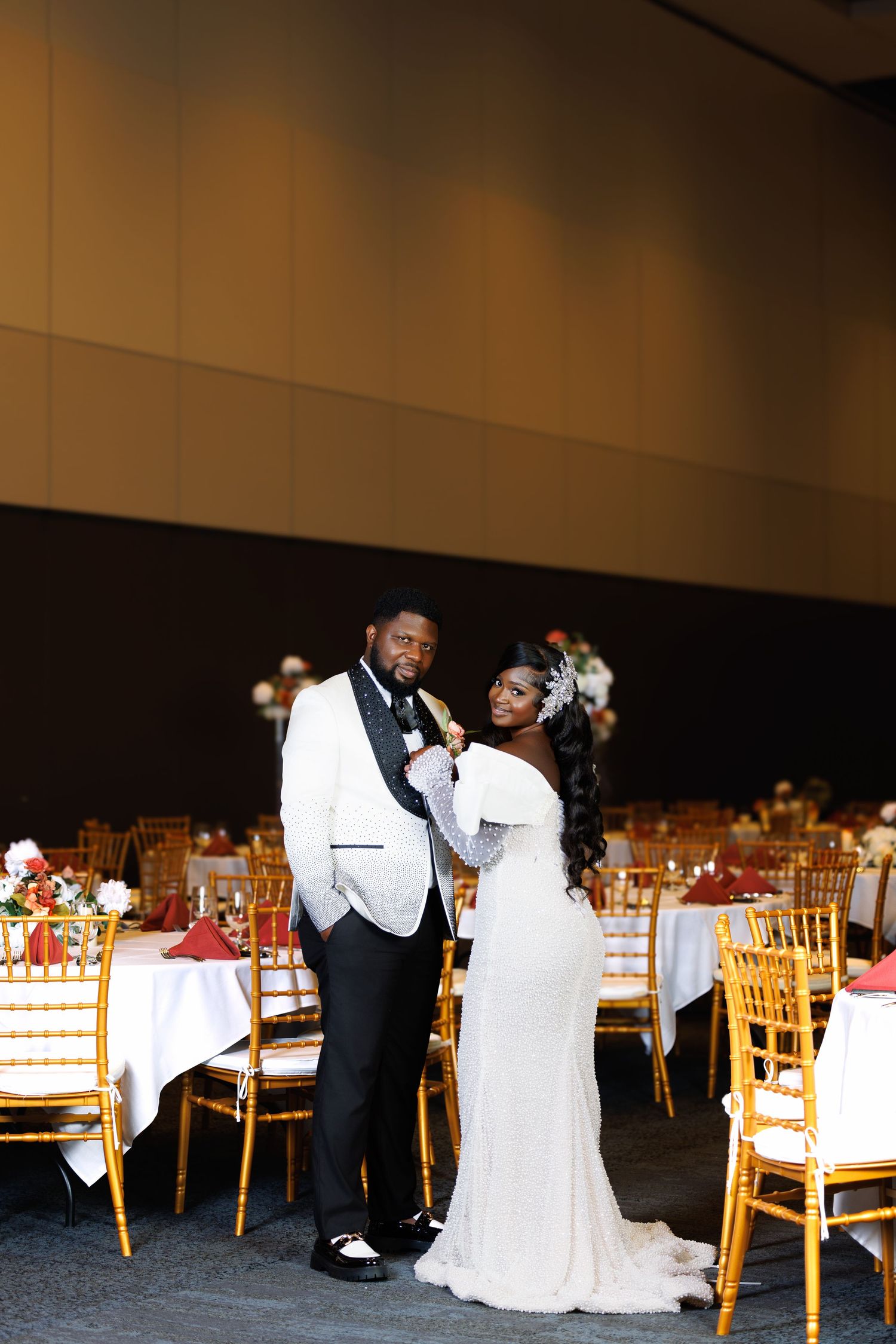 Elegant couple in formal wedding attire share a moment in an empty banquet hall with gold chairs and decorated tables.