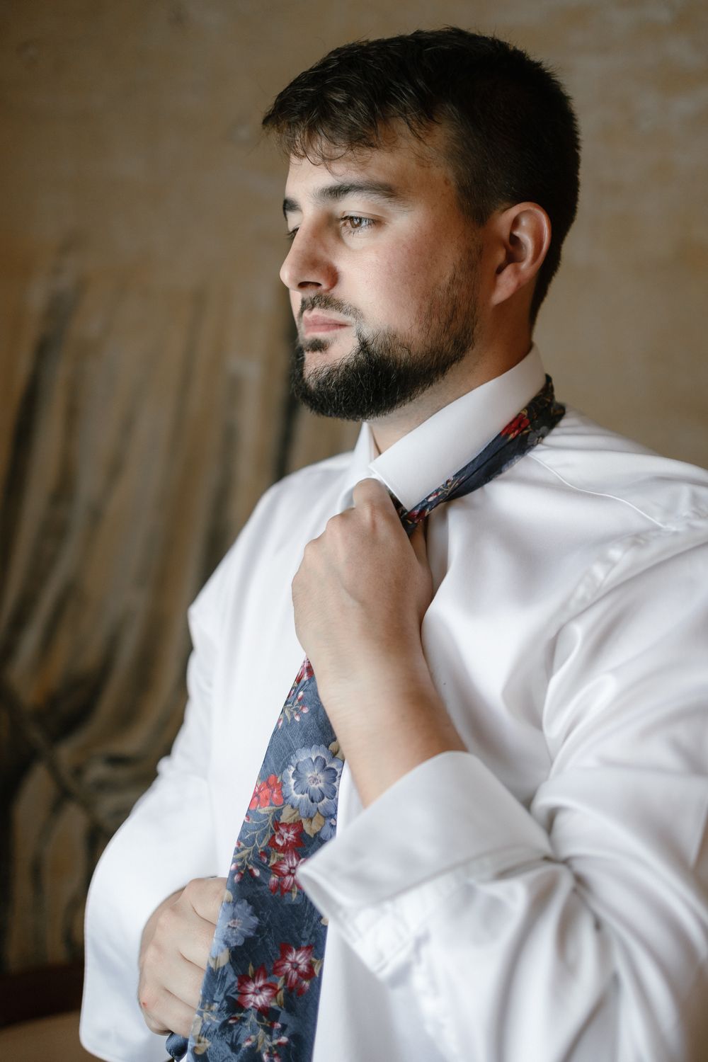 A man in a white dress shirt and floral tie adjusts his neckwear while looking to the side.