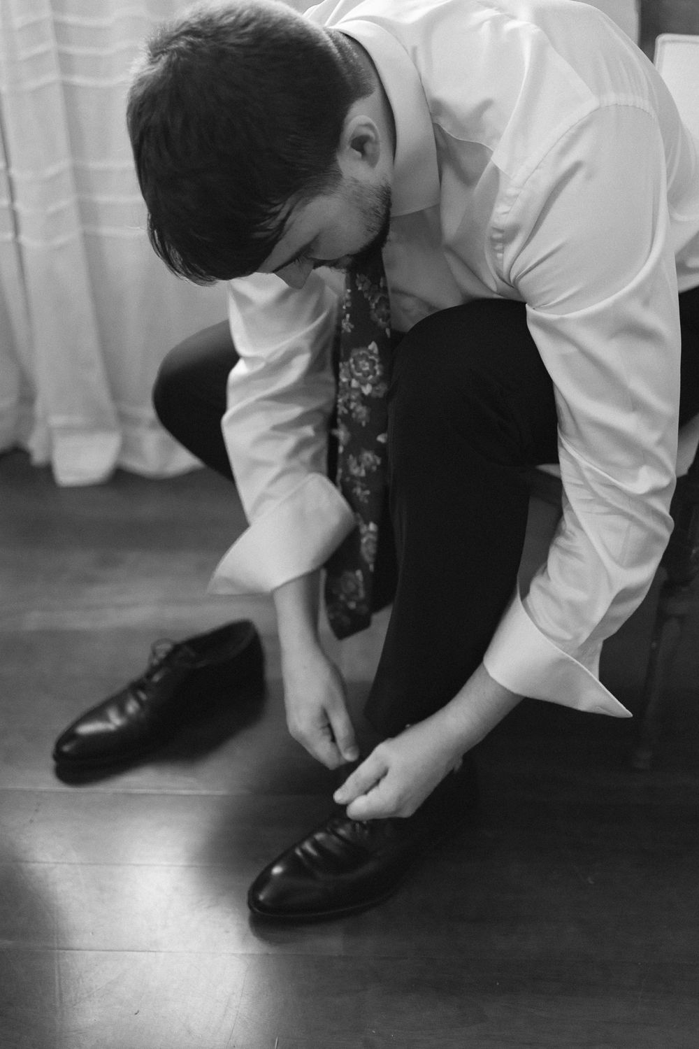 Person in formal attire bends down to tie their black dress shoes in a black and white image.