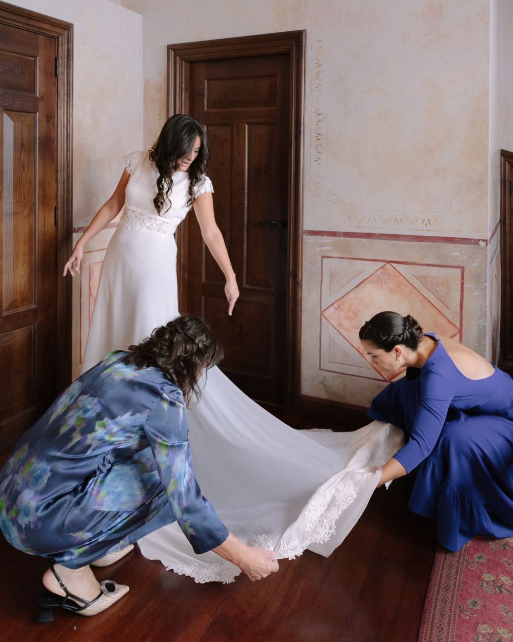 Two attendants help adjust a flowing white wedding dress train on a hardwood floor.