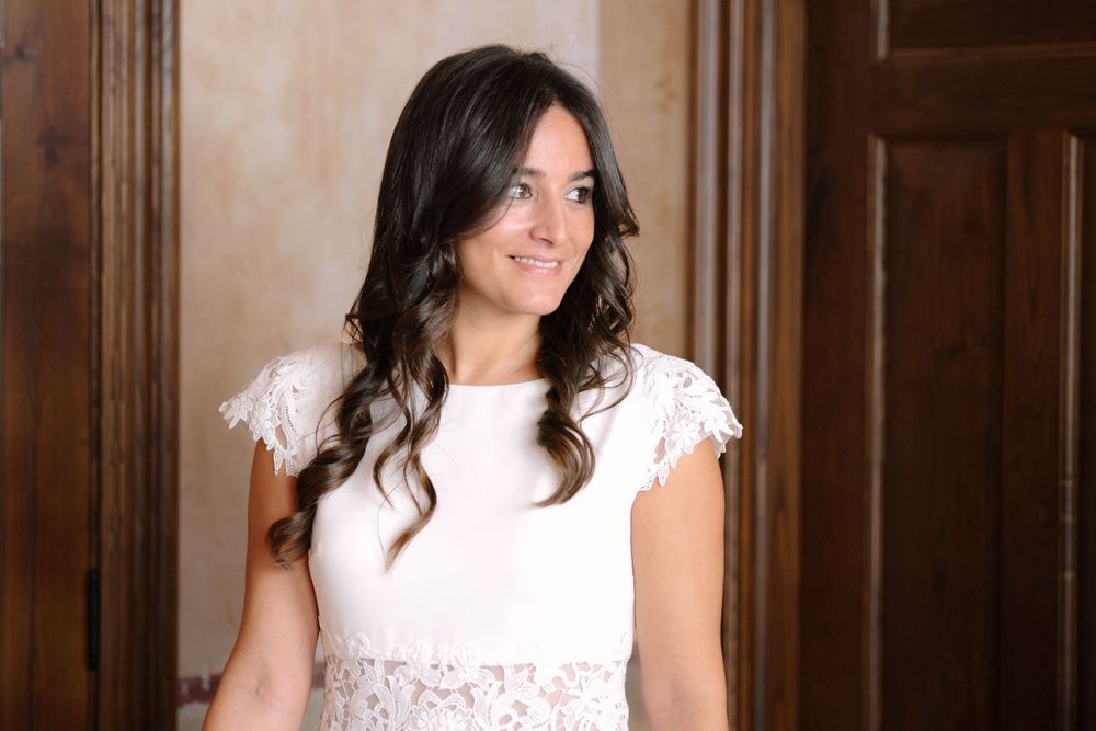 Woman in white lace dress with long dark wavy hair poses indoors against wooden wall.