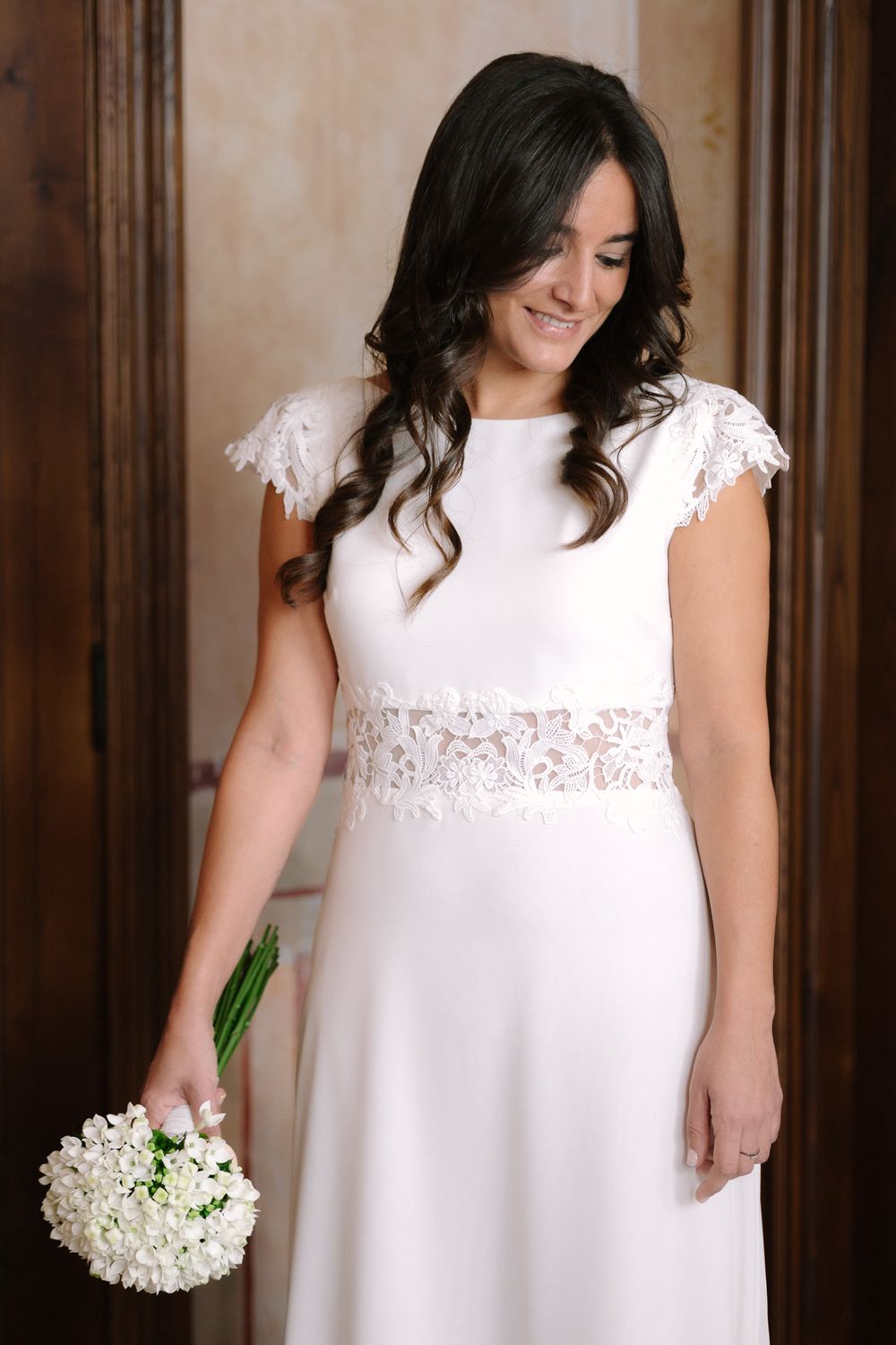 A white wedding dress with lace details and cap sleeves shown holding a small white bouquet.