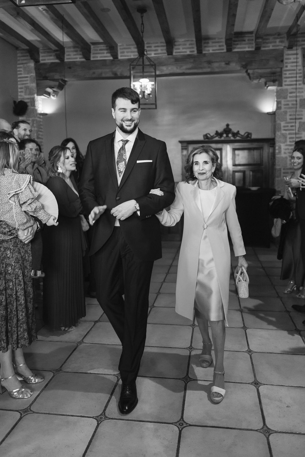 A formal black and white wedding procession photo showing a well-dressed man walking arm-in-arm with an older woman indoors.