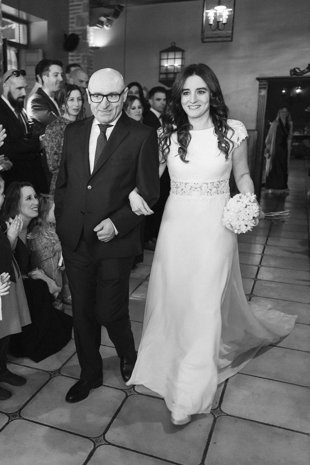 A formal wedding ceremony processional on decorative stone tiles with guests looking on.
